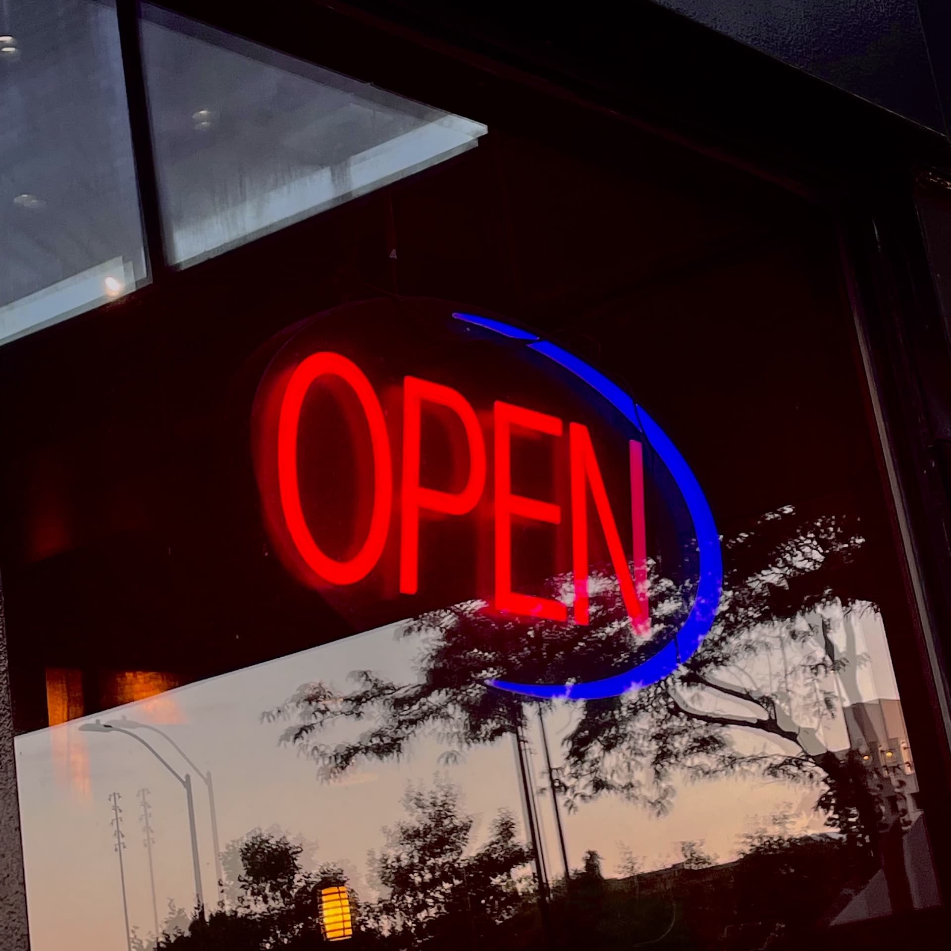 Reflection of a neon open sign, blurred trees, and the sunset