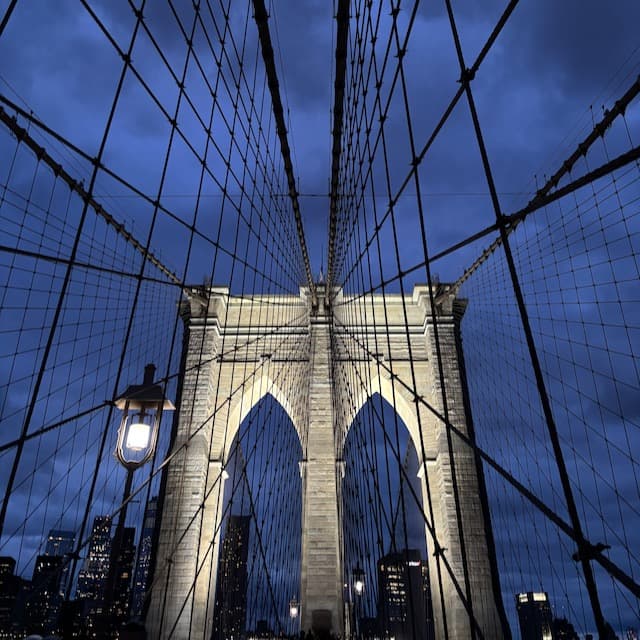 Brooklyn Bridge at dusk, view through cables. Illuminated stone tower against dark blue sky.