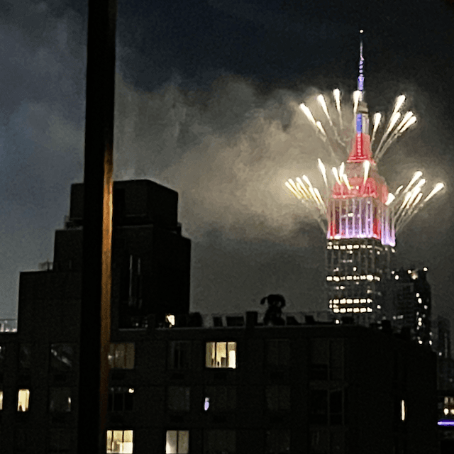 Dozens of fireworks bursting from the Empire State Building, decorated with red, white, and blue lights for July 4th