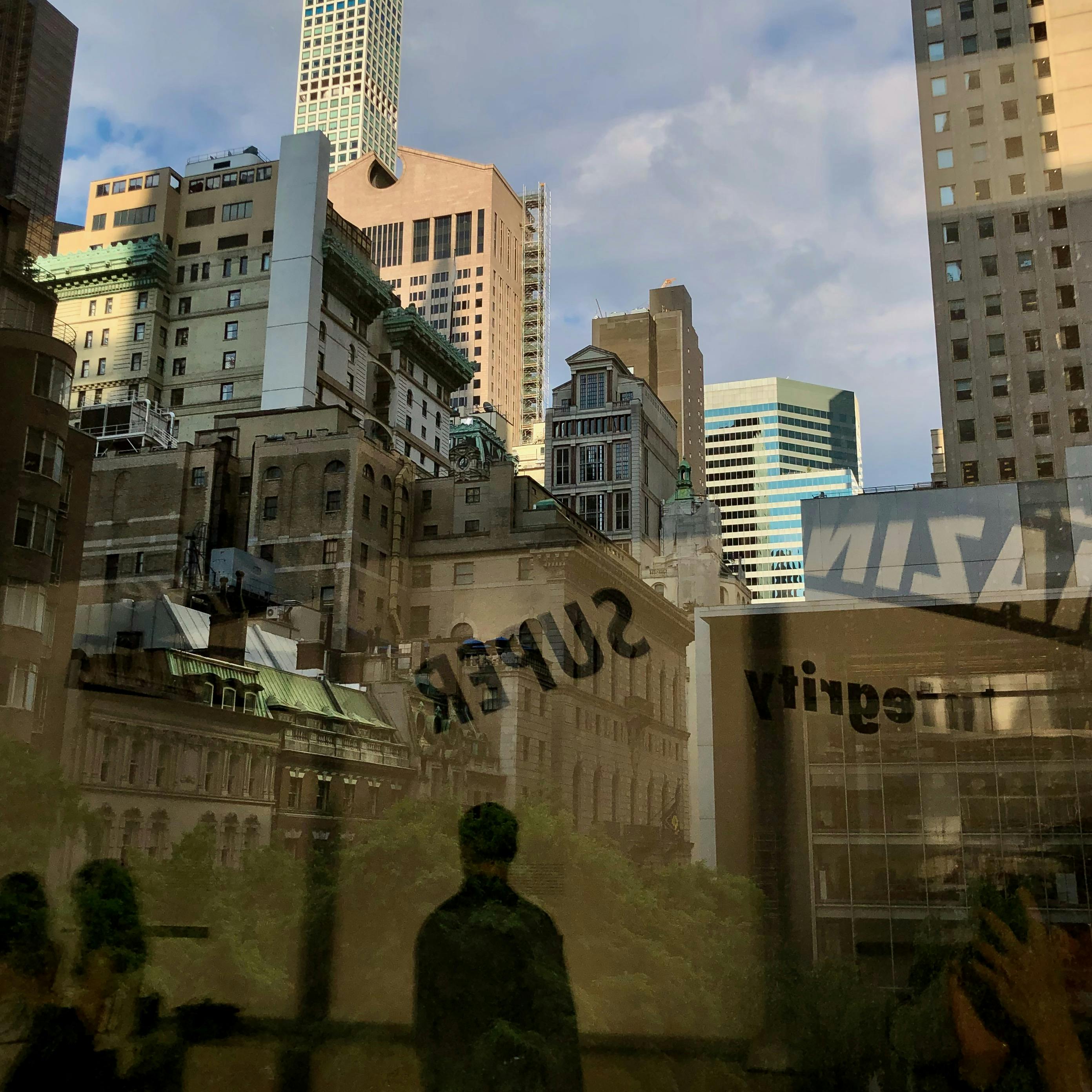 A dozen New York City skyscrapers viewed through a window at the MOMA featuring the shadowy reflections of three strangers