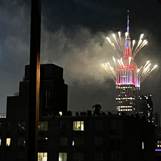 Dozens of fireworks bursting from the Empire State Building, decorated with red, white, and blue lights for July 4th