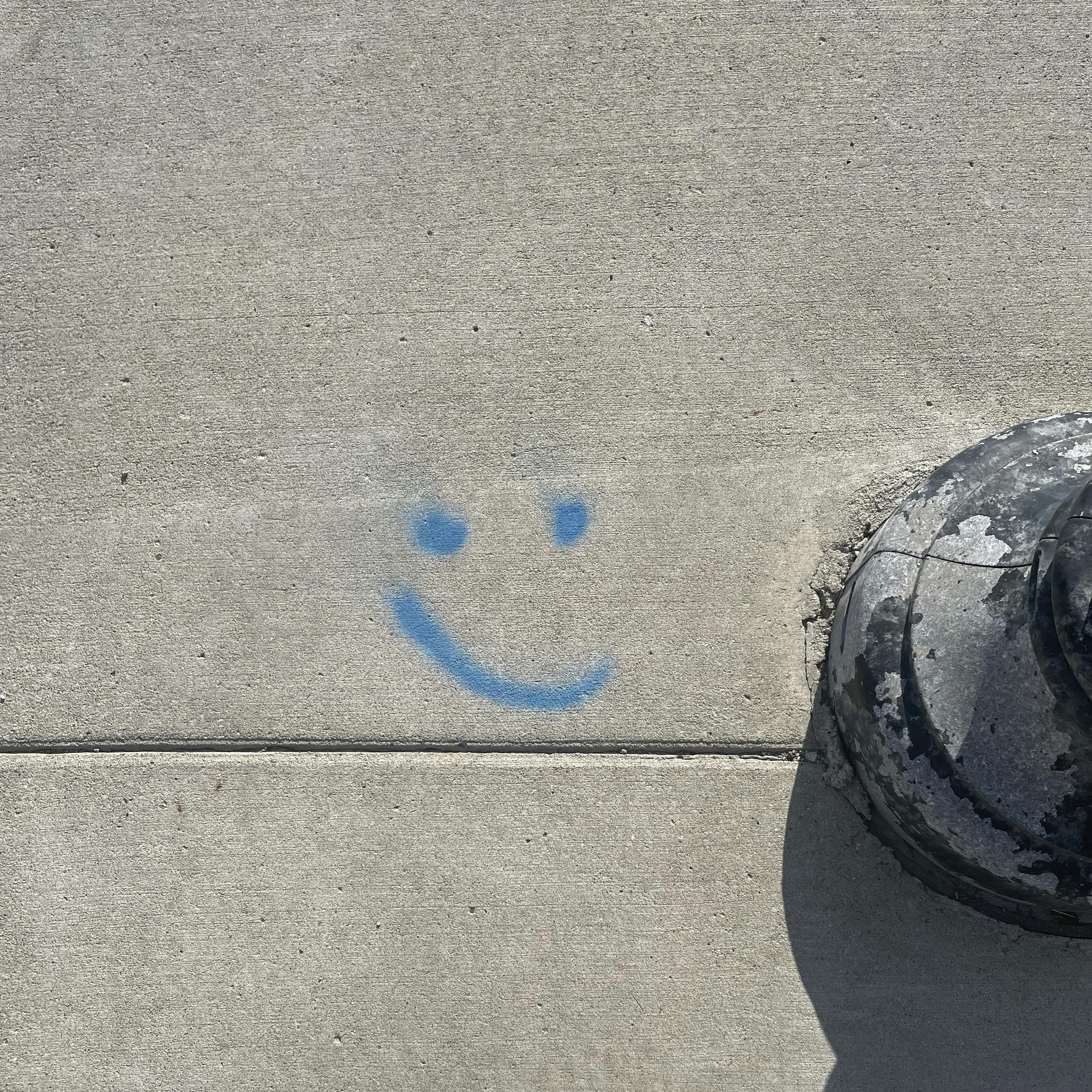 Aerial view of a blue smiley face with a crooked grin, spray painted on a sidewalk