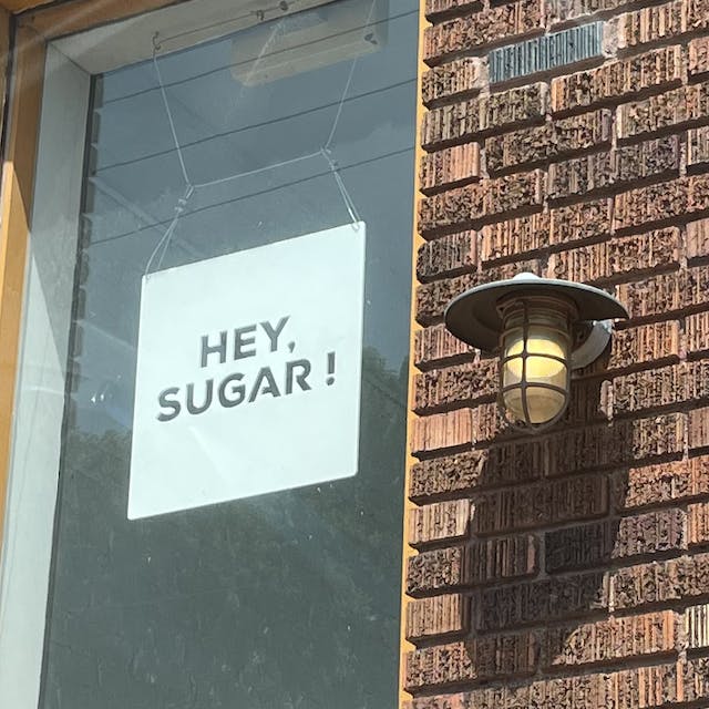 A sign in a window next to a brick wall that reads, 'HEY, SUGAR !'